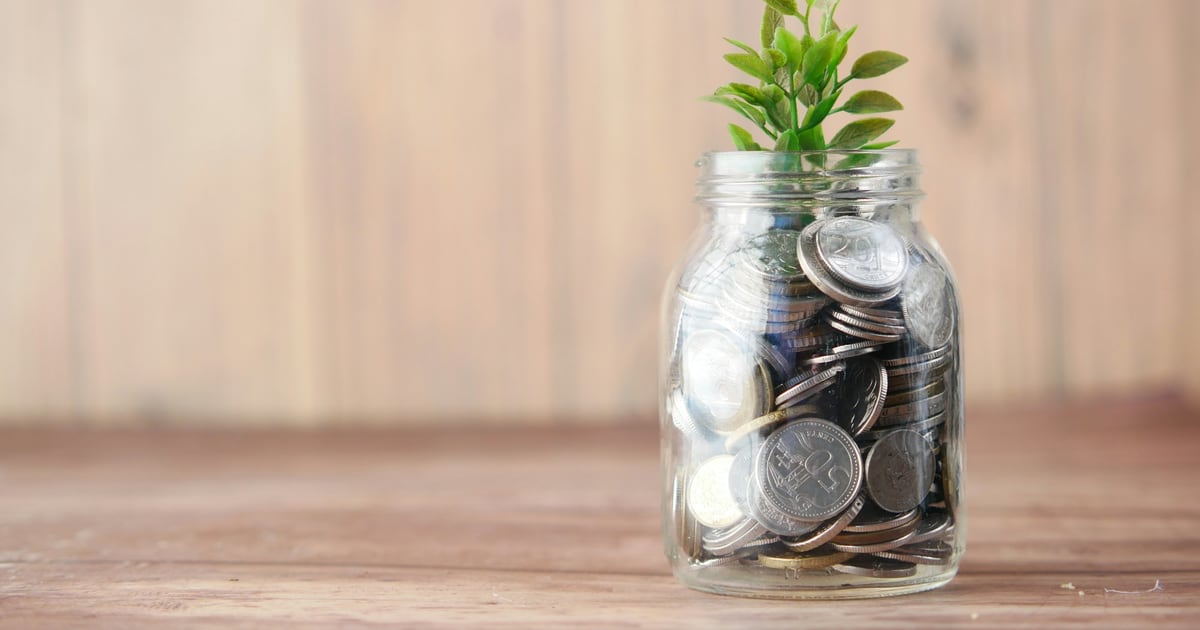 A glass jar of coins with a green seedling growing from the top, on a wooden table