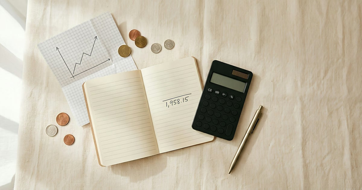 An open notebook with a written total, a calculator, a pen and a few coins on a linen cloth