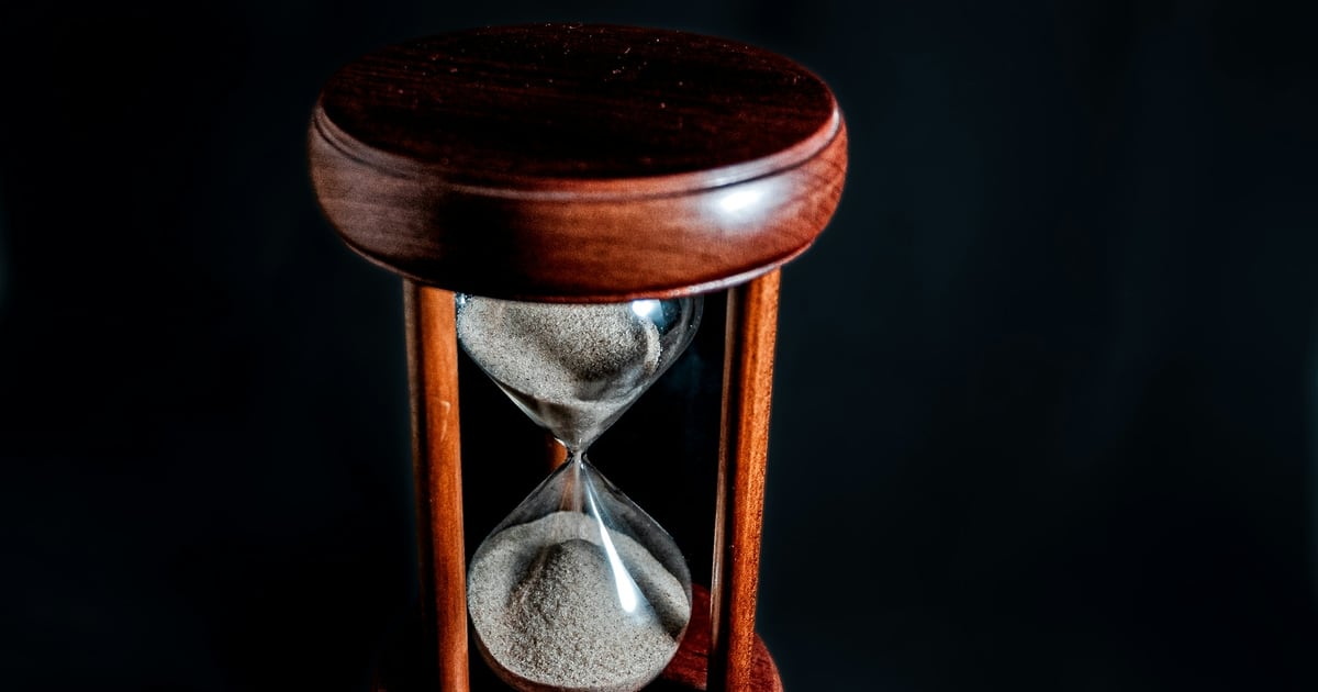 A wooden hourglass on a dark background with most of its sand still in the upper bulb