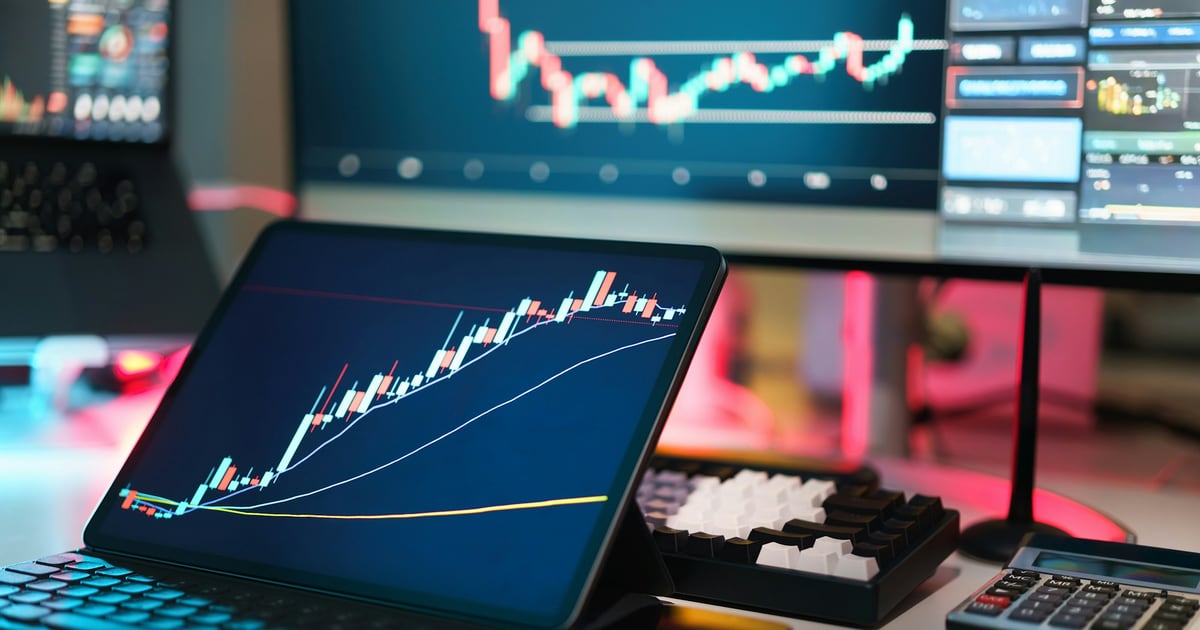 A tablet showing a rising candlestick chart, with trading screens and a keyboard behind it