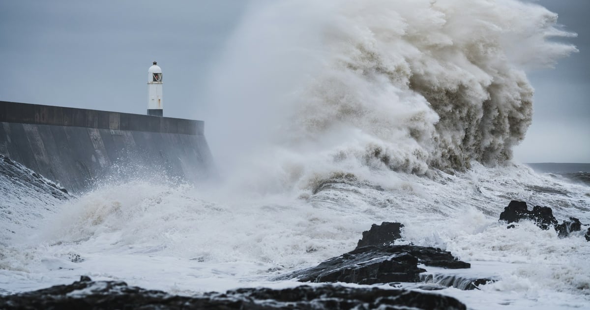 A storm wave breaking over a harbor wall beside a small white lighthouse